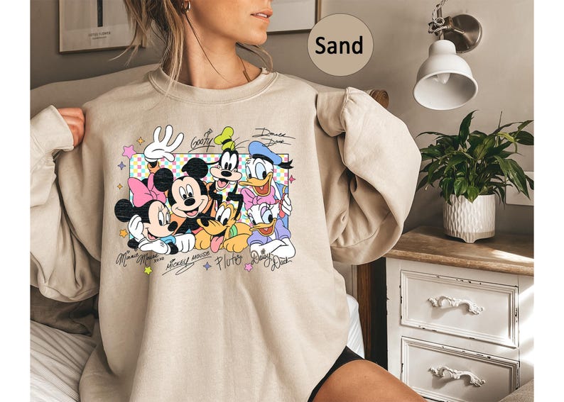 May include: Sand-colored sweatshirt featuring a colorful graphic of Disney characters including Mickey Mouse, Minnie Mouse, Goofy, Donald Duck, and Pluto. The design includes character signatures and a pastel-colored border.