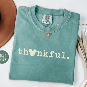 May include: Light green t-shirt with the word "thankful" in white, featuring a Mickey Mouse head in place of the "o". A gold necklace with a pendant rests on the shirt. A brown hat and dried plant are in the background.