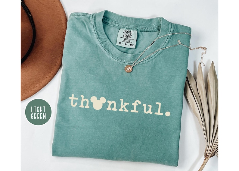 May include: Light green t-shirt with the word "thankful" in white, featuring a Mickey Mouse head in place of the "o". A gold necklace with a pendant rests on the shirt. A brown hat and dried plant are in the background.