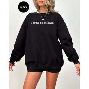 May include: Black sweatshirt with the text "I could be meaner" in white. The sweatshirt has a crew neck with a white collar and long sleeves. The model is wearing black shorts. The word "Black" is in a black circle in the top left corner.