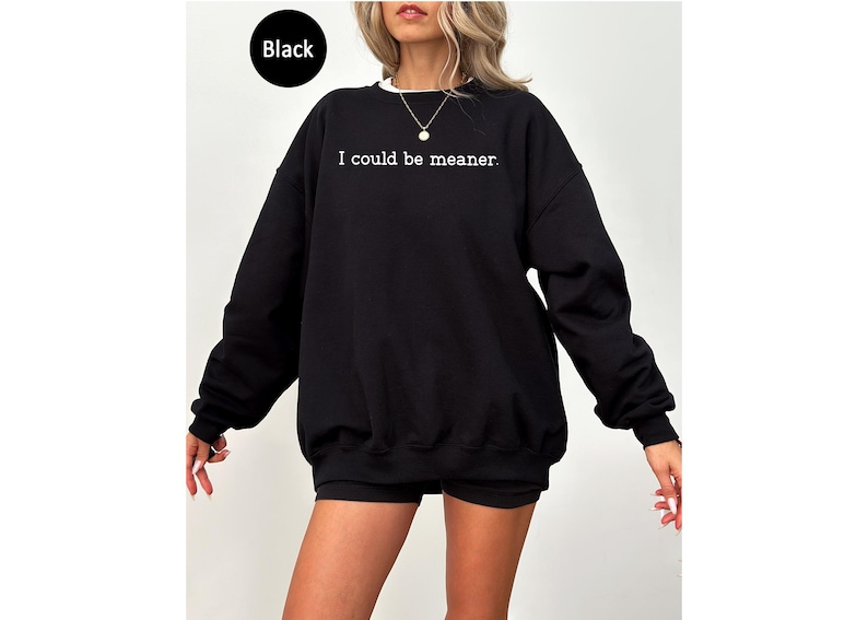 May include: Black sweatshirt with the text "I could be meaner" in white. The sweatshirt has a crew neck with a white collar and long sleeves. The model is wearing black shorts. The word "Black" is in a black circle in the top left corner.