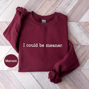 May include: Maroon crewneck sweatshirt with the text "I could be meaner" in white. The sweatshirt is folded on a light wood surface. A maroon circle with the word "Maroon" is in the lower left corner.