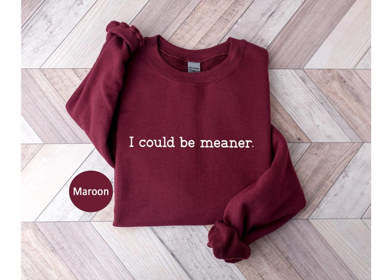May include: Maroon crewneck sweatshirt with the text "I could be meaner" in white. The sweatshirt is folded on a light wood surface. A maroon circle with the word "Maroon" is in the lower left corner.