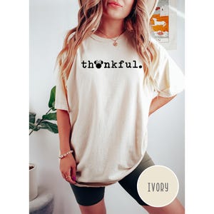May include: Ivory-colored t-shirt with the word "thankful" printed in black, featuring a Mickey Mouse head replacing the "o". The shirt is a relaxed fit, with short sleeves. The model is wearing dark green shorts.