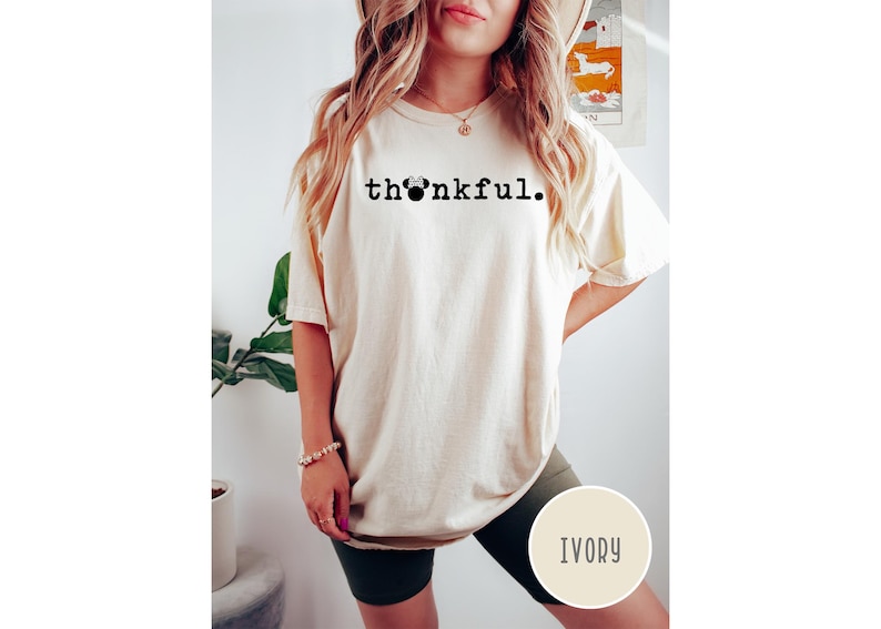 May include: Ivory-colored t-shirt with the word "thankful" printed in black, featuring a Mickey Mouse head replacing the "o". The shirt is a relaxed fit, with short sleeves. The model is wearing dark green shorts.