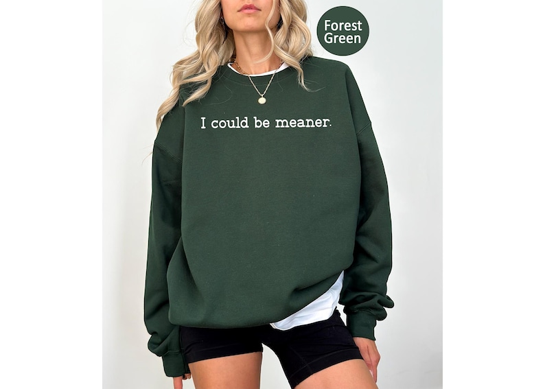 May include: Forest green sweatshirt with the text "I could be meaner." The long-sleeved crew neck sweatshirt has a white undershirt peeking out from the bottom. The sweatshirt is a solid color with a round green label in the upper right corner.