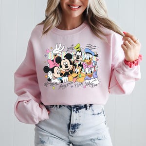 May include: Pink crewneck sweatshirt featuring a colorful graphic of Disney characters Mickey Mouse, Minnie Mouse, Goofy, Donald Duck, Daisy Duck, and Pluto. The characters' names are written below the image.