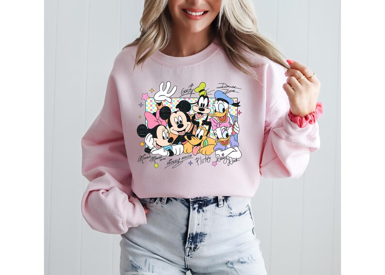 May include: Pink crewneck sweatshirt featuring a colorful graphic of Disney characters Mickey Mouse, Minnie Mouse, Goofy, Donald Duck, Daisy Duck, and Pluto. The characters' names are written below the image.