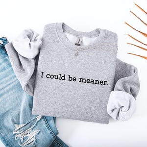 May include: A heather gray sweatshirt with the phrase "I could be meaner" printed in black. The sweatshirt is paired with light blue ripped jeans and a silver necklace. The sleeves are cuffed to reveal a white lining.