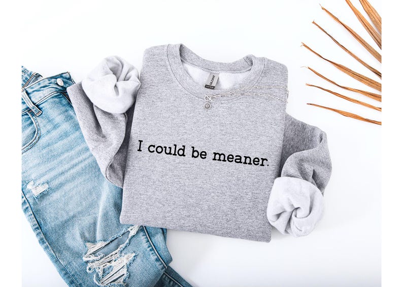 May include: A heather gray sweatshirt with the phrase "I could be meaner" printed in black. The sweatshirt is paired with light blue ripped jeans and a silver necklace. The sleeves are cuffed to reveal a white lining.
