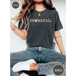 May include: Dark gray t-shirt with the word "thankful" in white, featuring a Mickey Mouse head in place of the "o". The shirt is paired with blue jeans and white sneakers. A casual, comfortable style.