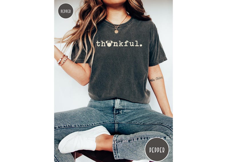 May include: Dark gray t-shirt with the word "thankful" in white, featuring a Mickey Mouse head in place of the "o". The shirt is paired with blue jeans and white sneakers. A casual, comfortable style.