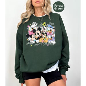 May include: Forest green sweatshirt featuring a graphic of Mickey Mouse, Minnie Mouse, Goofy, Donald Duck, and Pluto. The design includes the characters' signatures and colorful stars. The sweatshirt has a crew neck and long sleeves.