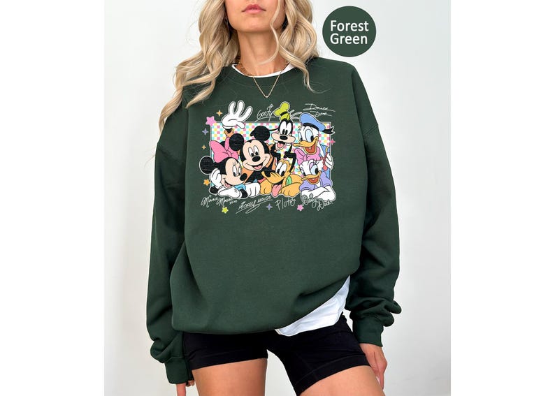 May include: Forest green sweatshirt featuring a graphic of Mickey Mouse, Minnie Mouse, Goofy, Donald Duck, and Pluto. The design includes the characters' signatures and colorful stars. The sweatshirt has a crew neck and long sleeves.