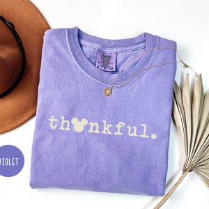 May include: A violet Comfort Colors t-shirt with the word "thankful" printed in light beige, featuring a Mickey Mouse head in place of the "o". A gold necklace and a brown hat are also visible.