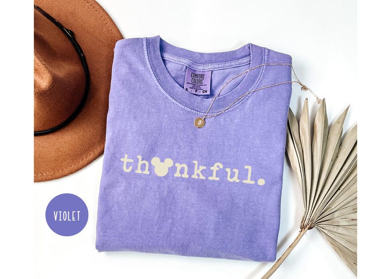 May include: A violet Comfort Colors t-shirt with the word "thankful" printed in light beige, featuring a Mickey Mouse head in place of the "o". A gold necklace and a brown hat are also visible.