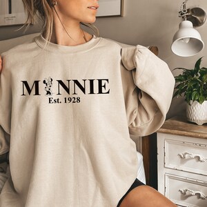 Pu&ograve; includere: Una felpa beige con una grafica nera di Minnie Mouse e il testo "MINNIE Est. 1928"