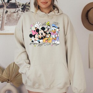 May include: Beige hoodie featuring a colorful graphic of Mickey Mouse, Minnie Mouse, Donald Duck, Daisy Duck, Goofy, and Pluto. The graphic includes their signatures and is set against a pastel background. The hoodie has a front pocket and a drawstring hood.