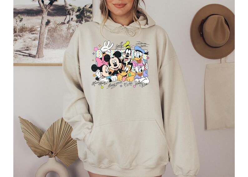 May include: Beige hoodie featuring a colorful graphic of Mickey Mouse, Minnie Mouse, Donald Duck, Daisy Duck, Goofy, and Pluto. The graphic includes their signatures and is set against a pastel background. The hoodie has a front pocket and a drawstring hood.
