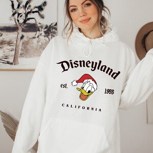 May include: White hooded sweatshirt with a black graphic print of Donald Duck wearing a Santa hat. The graphic also includes the text "Disneyland est. 1955 CALIFORNIA".