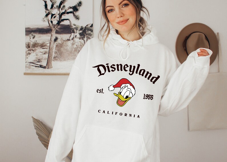 May include: White hooded sweatshirt with a black graphic print of Donald Duck wearing a Santa hat. The graphic also includes the text "Disneyland est. 1955 CALIFORNIA".