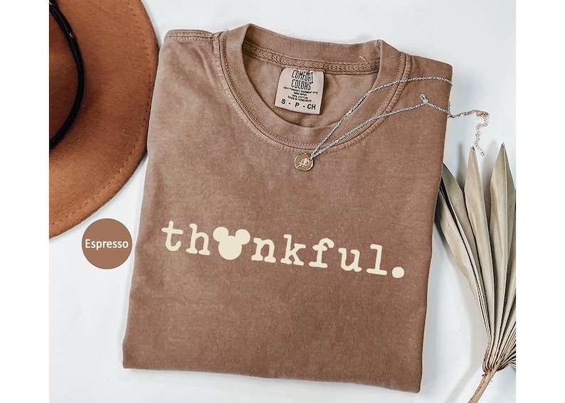 May include: A brown Comfort Colors t-shirt with the word "thankful" printed in white, featuring a Mickey Mouse head in place of the "o". A silver necklace with a small pendant rests on the shirt. A brown hat is visible in the upper left corner.