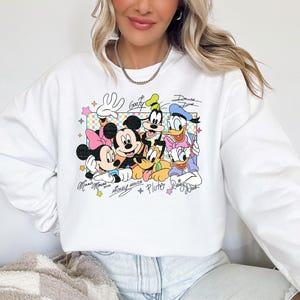 May include: White sweatshirt featuring a colorful graphic of Disney characters Mickey Mouse, Minnie Mouse, Donald Duck, Daisy Duck, Goofy, and Pluto. The design includes character signatures and a checkered background.