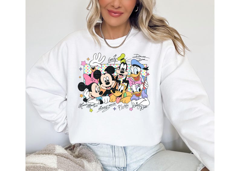 May include: White sweatshirt featuring a colorful graphic of Disney characters Mickey Mouse, Minnie Mouse, Donald Duck, Daisy Duck, Goofy, and Pluto. The design includes character signatures and a checkered background.