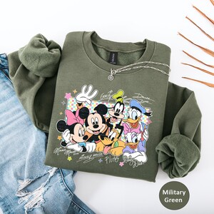May include: Military green sweatshirt featuring a graphic of Mickey Mouse, Minnie Mouse, Goofy, Donald Duck, and Pluto. The design includes character signatures and colorful accents. A silver necklace is draped on the sweatshirt, which is paired with distressed blue jeans.