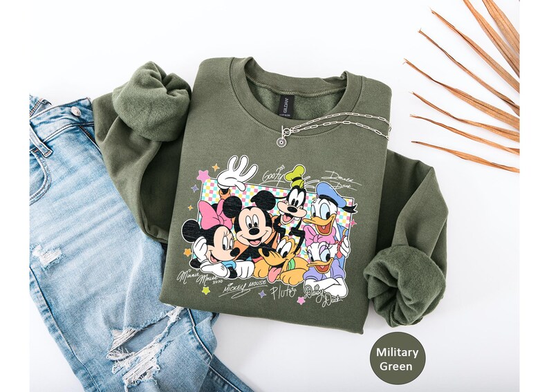 May include: Military green sweatshirt featuring a graphic of Mickey Mouse, Minnie Mouse, Goofy, Donald Duck, and Pluto. The design includes character signatures and colorful accents. A silver necklace is draped on the sweatshirt, which is paired with distressed blue jeans.