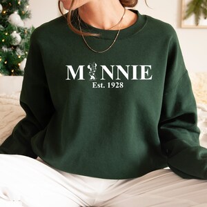 Pu&ograve; includere: Una felpa verde scuro con una grafica bianca di Minnie Mouse e il testo "Minnie Est. 1928".