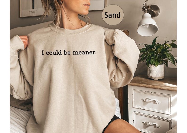 May include: Sand-colored sweatshirt with the text "I could be meaner" in black. The long-sleeved top is a neutral color and has a relaxed fit. The sweatshirt is part of a clothing collection.