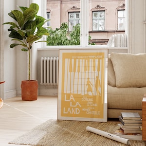 Peut inclure: Une affiche jaune avec le texte "La La Land" en lettres blanches. L'affiche représente une silhouette de couple dansant sur un clavier de piano. L'affiche est accrochée à un mur blanc dans un salon.