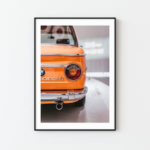 May include: A close-up of the rear end of an orange vintage BMW 2002ti car. The car has a chrome bumper and a single exhaust pipe.