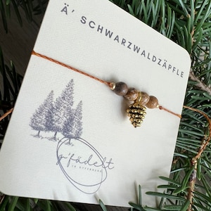Könnte beinhalten: Ein braunes und goldenes Armband mit einem Tannenzapfen-Anhänger. Das Armband ist aus natürlichen Holzperlen und einer braunen Schnur gefertigt. Die Karte hinter dem Armband lautet "Ä' SCHWARZWALDÄPFLE" und "gfädelt in offenburg".