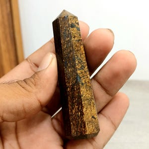 Op de afbeelding: Een bruine en zwarte edelsteen obelisk met een puntige top. De steen heeft een textuur met een mix van donkerbruine en goudbruine tinten. De obelisk wordt in een hand gehouden.