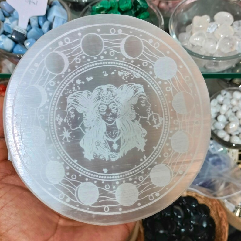 May include: A circular selenite crystal plate with an etched design. The design features a central image of a three-faced woman surrounded by moon phases and stars. The plate is held in a hand.