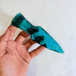Puede incluir: Una astilla de vidrio de color turquesa, de forma irregular y con un extremo puntiagudo. El vidrio translúcido presenta zonas más oscuras en su interior. El objeto se sostiene en una mano, mostrando su tamaño y forma.