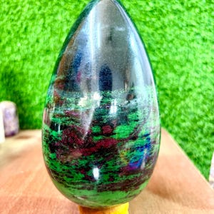 Puede incluir: Una piedra pulida en forma de huevo con una mezcla de colores verdes, rojo oscuro y negro. La piedra está montada sobre un pequeño soporte redondo de madera. El fondo es un césped artificial verde brillante.