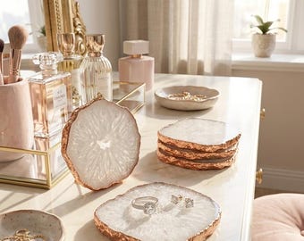 May include: A set of white agate coasters with metallic copper-toned edges. The coasters are circular and square in shape, featuring a crystalline centre texture. One square coaster holds a silver ring and two stud earrings, while others are stacked.