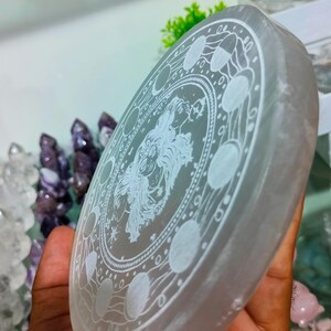 May include: A circular selenite crystal plate with an etched design. The design features a central figure surrounded by phases of the moon and celestial symbols. The plate is held in a hand, with other crystals visible in the background.