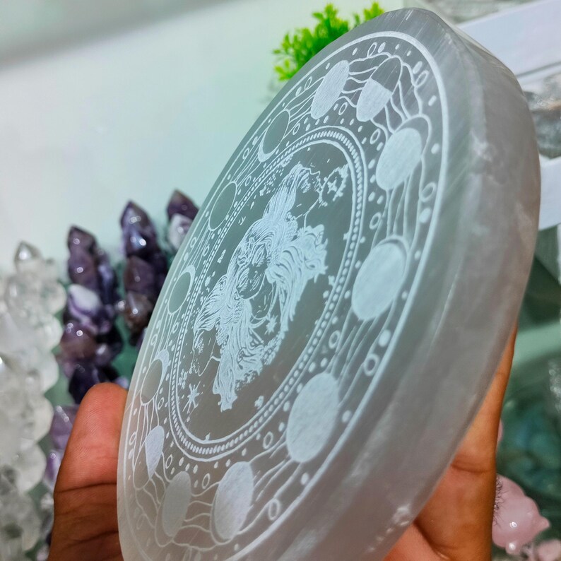 May include: A circular selenite crystal plate with an etched design. The design features a central figure surrounded by phases of the moon and celestial symbols. The plate is held in a hand, with other crystals visible in the background.