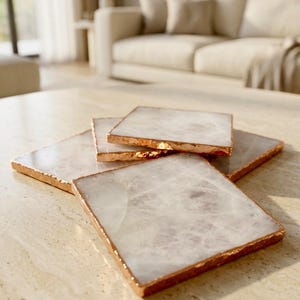 May include: A set of four square coasters made of translucent white stone with a polished finish. Each coaster features a metallic copper-colored edge that highlights the natural texture and irregular shape of the stone's perimeter.