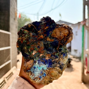 May include: A rough, natural azurite and malachite specimen. The stone features a mix of deep blue, turquoise, green, and brown hues. The mineral has a textured, uneven surface, showcasing its raw, unpolished state. A unique piece for collectors.