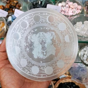 May include: A circular selenite crystal plate with an etched design. The design features a three-faced goddess surrounded by moon phases. The plate is white and held in a hand, with other crystals visible in the background.