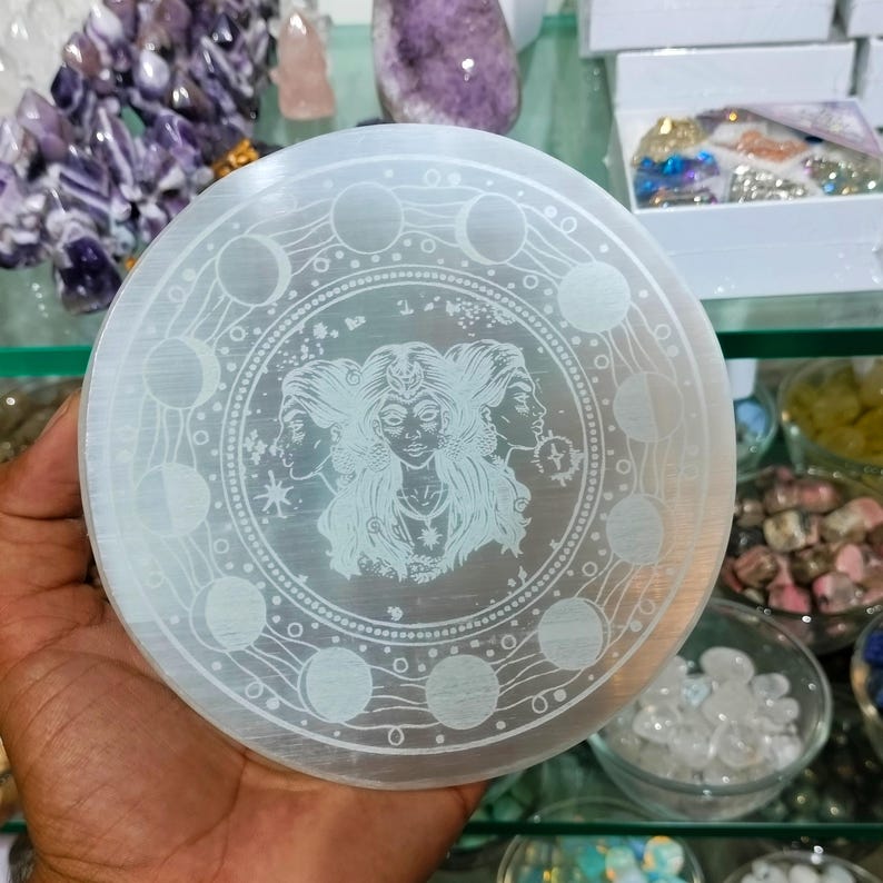 May include: A circular selenite plate with an etched design. The design features three female faces in the center, surrounded by a circle of moon phases. The plate is held in a hand, with other crystals and gemstones visible in the background.