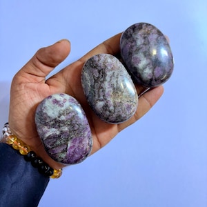 May include: Three polished, oval-shaped stones with a mix of purple, gray, and white hues. The stones are smooth and appear to be natural gemstones, held in a hand against a light blue background.