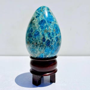 Puede incluir: Una piedra pulida en forma de huevo, en tonos azules y turquesas, exhibida sobre un soporte de madera oscura. La superficie de la piedra presenta un patrón moteado. El soporte es de color marrón oscuro, con un diseño clásico. La estética general es elegante y natural.