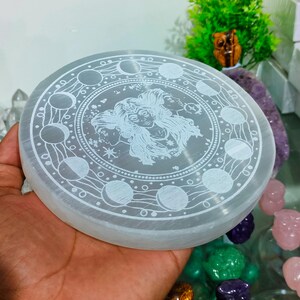 May include: A round, white selenite crystal plate with an etched design. The design features a woman's face surrounded by moon phases and celestial elements. The plate is approximately 10 cm in diameter. A spiritual or decorative item.