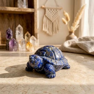 May include: A detailed, blue lapis lazuli turtle figurine. The sculpture has a textured shell and detailed legs and head. The figurine is on a light-colored surface, with crystals and decorative items in the background.
